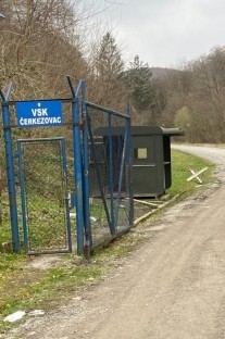 Seosko područje s makadamskom cestom koja vodi do ograđene kapije s natpisom "VŠK Čerkezovac" u plavoj i bijeloj boji. Iza vrata nalazi se mala građevina tamne boje u blizini drveća i brda u pozadini.
